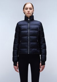 Zwarte pufferjack met hoge kraag, horizontaal gestikt patroon, ritssluiting aan de voorkant en zijzakken; gecombineerd met aansluitende zwarte leggings.
