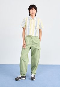 Gekürztes, gelb-blau gestreiftes Hemd mit zwei Brusttaschen, kombiniert mit lockeren hellgrünen Cargo-Hosen und grauen Sneakers mit weißen Akzenten.