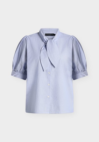 Blouse bleu clair à manches courtes bouffantes, boutonnée à l’avant, avec un col foulard noué.