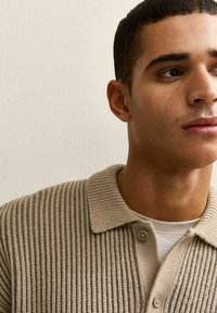 Młody mężczyzna z krótkimi włosami, ubrany w beżowy prążkowany kardigan na białej koszuli, patrzący w prawo na tle gładkiej ściany.