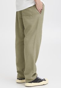 Olive green wide-leg pants with a textured finish, featuring side pockets and a classic fit, paired with black shoes with white laces.