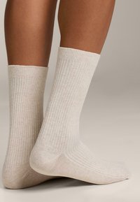 Piedi che indossano calze a costine beige chiaro fino al polpaccio, su uno sfondo neutro, evidenziando la texture e la vestibilità delle calze sulla pelle.