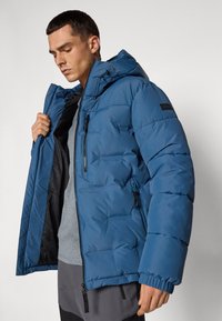 Chaqueta azul acolchada con capucha, que cuenta con bolsillos con cremallera, puños elásticos y una textura suave. El forro interior es negro.