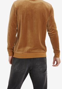 Sudadera larga de terciopelo marrón con cuello redondo y puños acanalados, combinada con unos vaqueros oscuros que tienen dos bolsillos traseros.