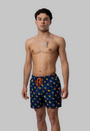 Giovane uomo in piedi, senza maglietta, indossa pantaloncini da bagno blu navy con motivi colorati e coulisse arancione, sfondo grigio neutro.