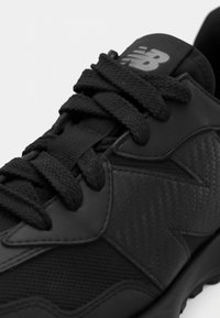 Kratki prikaz crne New Balance tenisice s teksturiranim sintetičkim materijalom i debelim, okruglim vezicama zavezanima na vrhu.
