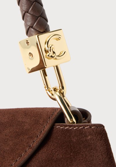 Gros plan d’une boucle logo dorée et d’une sangle tressée attachées à un sac à main en daim marron.
