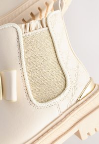 Bottine couleur crème avec un panneau latéral élastique texturé, des accents tissés et une semelle en caoutchouc robuste à motif léger.