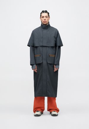 Long manteau gris avec une partie supérieure raccourcie, deux poches avant avec des fermetures éclair orange, manches à plusieurs couches et un col droit. Porté avec un pantalon orange.
