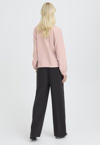 Woman with long blonde hair wearing a light pink long-sleeve blouse and black wide-leg trousers, standing facing away on a plain background.