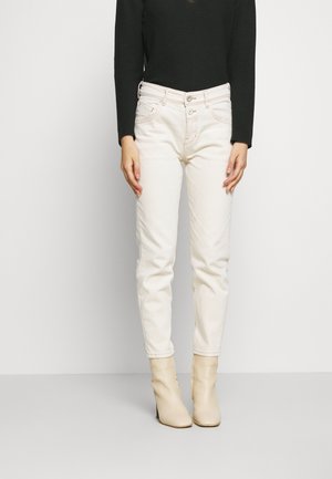 Personne portant un jean blanc taille haute avec fermeture à double bouton, un haut noir à manches longues et des bottines beiges, debout devant un fond uni.