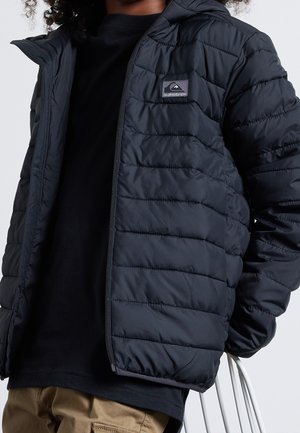 Person iført en sort quiltet lynlåstrøje med et logoemblem på brystet, sort skjorte under og beige bukser, der sidder på en hvid stol.