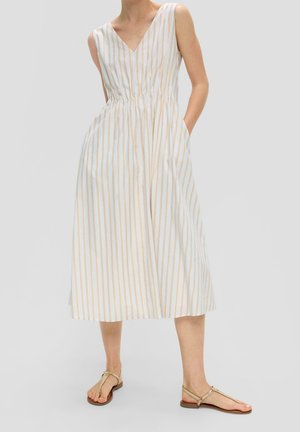 Frau trägt ärmelloses weißes Kleid mit vertikalen beigen Streifen und Taschen, kombiniert mit beigen flachen Zehensandalen.