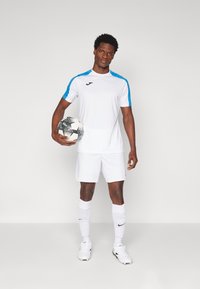 Hvitt idrettsjersey med blå detaljer, kombinert med hvite shortser og svarte fotballsko. Holder en svart og hvit fotball.