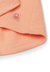 Licht perzikkleurige stof met een ribbelstructuur, afgeronde zoom en een vierkante merklabel met een logo in dezelfde kleur gestikt.