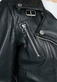 Černá kožená moto bunda s texturovaným povrchem, vybavená stříbrnými kovovými doplňky, diagonálním zipovým kapsou a knoflíkovými detaily na límci.