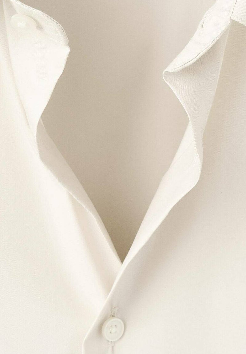 Chemise blanche boutonnée avec une texture douce, dotée d'un col pointu et d'un détail de bouton unique au niveau du cou, partiellement ouverte.