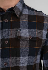 Camisa de franela a cuadros en negro, gris y naranja. Presenta cierre de botones, cuello, bolsillo en el pecho y textura de tejido tejido.