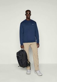 Marine genser, beige bukser, svart ryggsekk, hvite sneakers og solbriller. Genseren har logo på brystet, ryggsekken har flere rom.