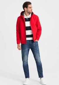Rød zip-opp jakke med hætte, parret med en hvid og navy-stribet skjorte, blå jeans og hvide sneakers. Glat stof, atletisk pasform.