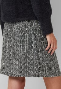 Nahaufnahme einer Person, die einen strukturierten Rock mit schwarz-weißer Musterung und ein flauschiges schwarzes Langarmoberteil trägt, mit der Hand entspannt an ihrer Seite.