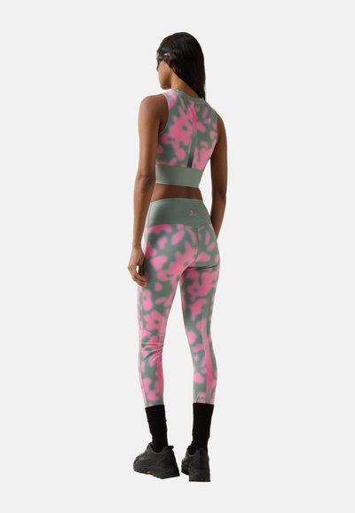 Frau trägt grünes und pinkes, camouflagegemustertes ärmelloses Crop-Top und passende Leggings, schwarze Socken und dunkle Sneakers, steht mit dem Rücken zur Kamera.