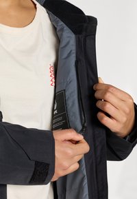 Černá bunda se šedým interiérem. Obsahuje kapsu na zip a manžetu na suchý zip. Pod ní je jednobarevné béžové tričko s červeným designem.