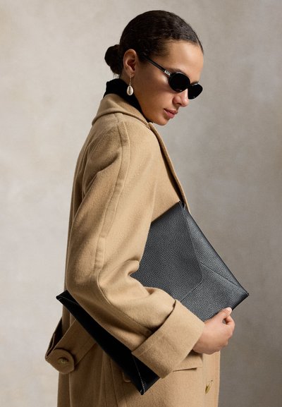 Frau mit beigem Mantel und schwarzen Sonnenbrillen hält eine schwarze, strukturierte Clutch vor einem schlichten neutralen Hintergrund.
