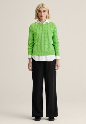 Ung kvinde med kort blondt hår iført en limegrøn kabelstrikket sweater over en hvid skjorte med krave og sorte vidde bukser.