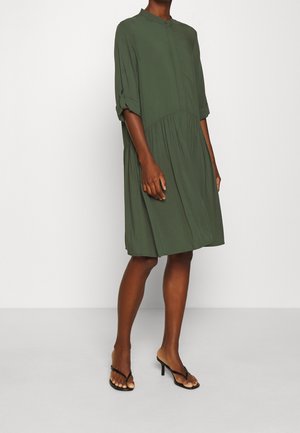 Femme portant une robe ample olive verte longueur genou avec des manches retroussées aux trois quarts et des sandales noires à talons bas, debout devant un fond uni.