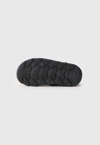 Schwarze Gummisohle mit einem strukturierten Profilmuster, das aus Winkelformen besteht, und einem runden Logo auf der Unterseite für Grip und Traktion.