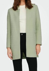 Abrigo de espiga de color verde claro con corte recto, frente abierto y mangas largas, combinado con una blusa color crema y pantalones negros.