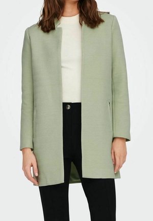 Cappotto herringbone verde chiaro con taglio dritto, apertura frontale e maniche lunghe, abbinato a un top crema e pantaloni neri.