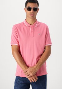 Lys rosa polo-skjorte med hvitstripete krage og mansjetter. Har to knapper og en liten logo på brystet. Kombinert med mørk denim.
