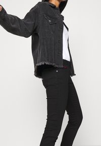 Chaqueta de mezclilla negra con un dobladillo deshilachado, dos bolsillos en el pecho y un corte relajado, combinada con pantalones negros ajustados que presentan sutiles detalles de costura.