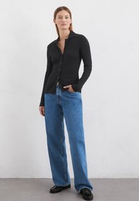 Schwarzes, geripptes, langärmliges Button-Up Hemd, kombiniert mit hellblauen, weiten Jeans und schwarzen Schuhen, vor einem neutralen Hintergrund.