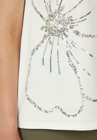 Witte shirt met een bloemenontwerp van sequins in verschillende maten in zilver. De sequins creëren een textuur met een sprankelend effect.
