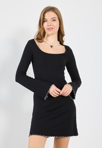 Abito nero a coste con maniche lunghe a sbuffo, dotato di bordo bianco a scallop alle maniche e all'orlo. Scollo quadrato e design aderente.