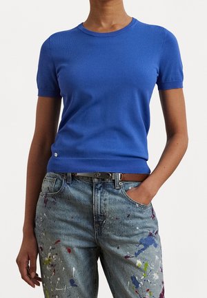 Femme portant un pull bleu ajusté à manches courtes, rentré dans un jean taille haute avec des éclaboussures de peinture colorées et une ceinture marron.