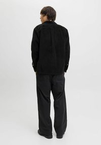 Une personne se tenant de dos, portant une veste en velours côtelé noir, un pantalon large noir et des chaussures noires, devant un fond clair uni.