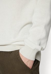 Jasnoszary dziergany sweter z prążkowanymi mankietami, zestawiony z brązowymi, w kratę spodniami. Faktura wydaje się miękka i przewiewna.