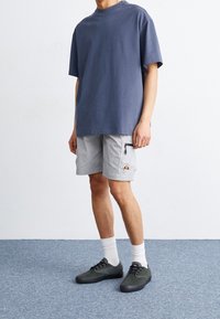 Persona con una camiseta azul marino holgada, pantalones cortos de cargo grises claros con un bolsillo con cremallera, calcetines blancos y zapatillas oscuras verdes, de pie sobre una alfombra azul.