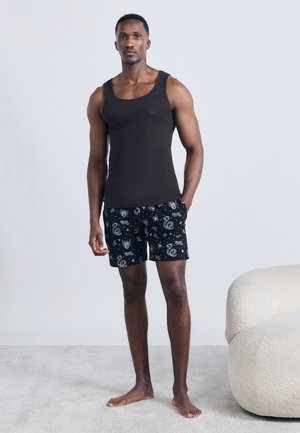 Hombre descalzo de pie, vistiendo una camiseta sin mangas negra y pantalones cortos negros con patrones de animales y temática espacial en una habitación minimalista.