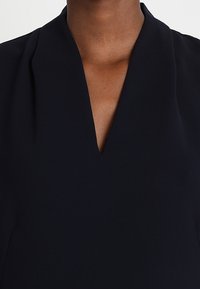Close-up of a dark-skinned person wearing a black top with a deep V-neck collar on a white background.