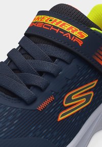 Σκούρος μπλε αθλητικός παπούτσι Skechers με μαύρα κορδόνια, πορτοκαλί και κίτρινο λογότυπο Skechers και φωτεινό κίτρινο επενδεδυμένο γιακά ορατό.