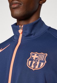 Navy athletic jacket with a high collar, orange zipper, and FC Barcelona logo. The fabric is smooth with a fitted design.