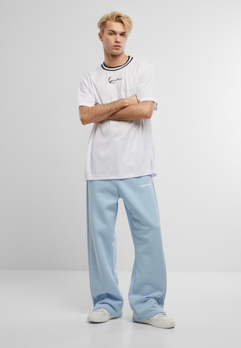 Bărbat blond stă cu brațele încrucișate, purtând un tricou alb și pantaloni largi de trening bleu, cu pantofi sport albi, pe un fundal simplu.