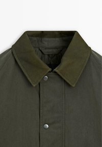 Olijfgroene jas met een matte afwerking, voorzien van een zachte textuur, drukknoopsluiting en een contrasterende kraag. Opvallend detail: raglanmouwen.