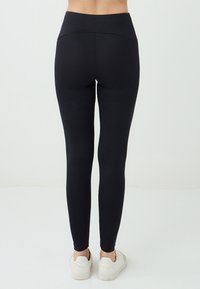 Una persona con leggings neri e sneakers bianche è rivolta di spalle, su uno sfondo chiaro, mostrando la vestibilità dell'abbigliamento.