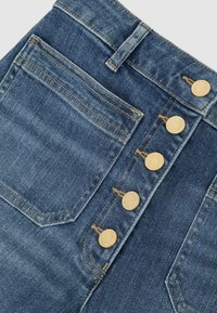 Prim-plan cu blugi din denim albastru, arătând buzunarul frontal, bucla pentru curea și cinci nasturi metalici aurii la fermoar, cu cusături portocalii.